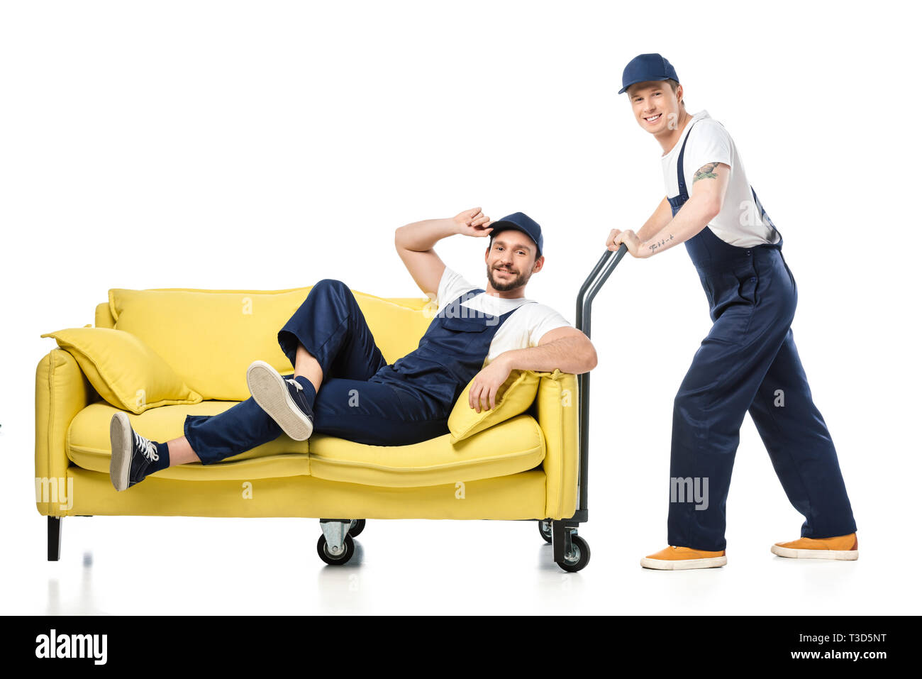 Mover in Uniform sitzen auf einem Sofa und Blick auf Kamera, während Kollege drücken Hand-Lkw auf Weiß Stockfoto