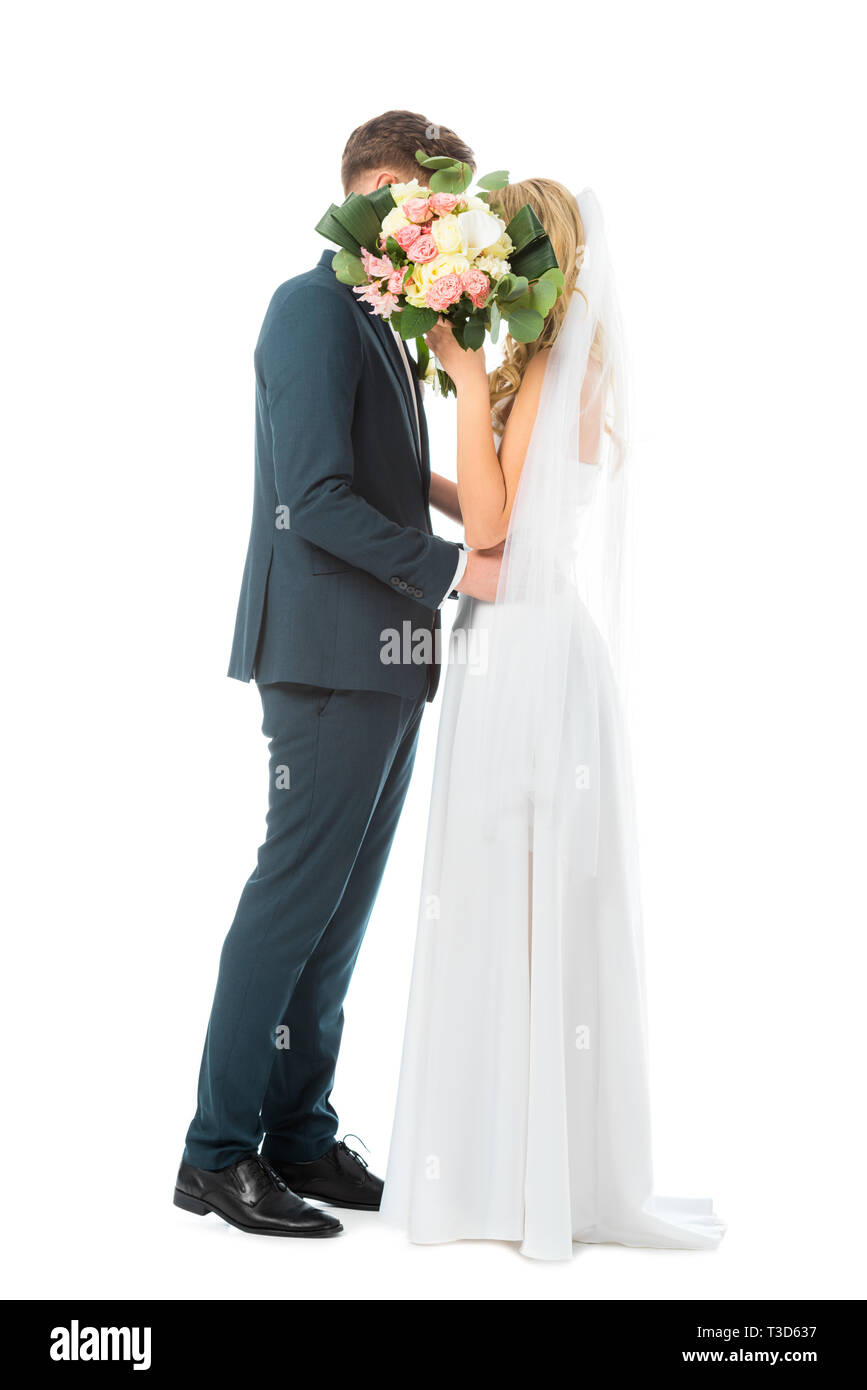 Braut im Brautkleid und Bräutigam im eleganten Anzug umarmen beim Verstecken Gesichter hinter wedding bouquet isoliert auf weißem Stockfoto