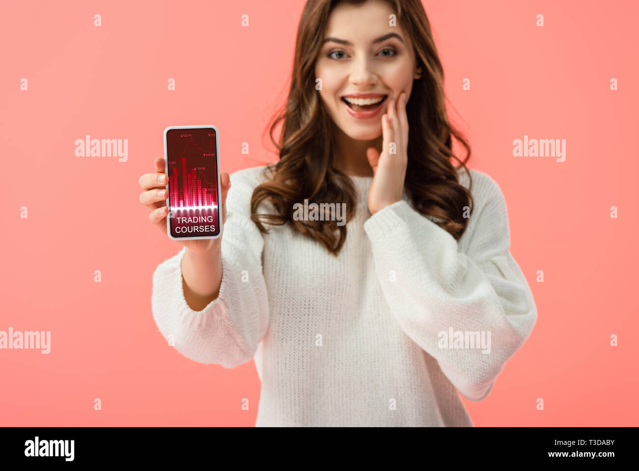 Selektiver Fokus der Frau in weißem Pullover holding Smartphone mit Handel Kurse App auf dem Bildschirm isoliert auf Rosa Stockfoto