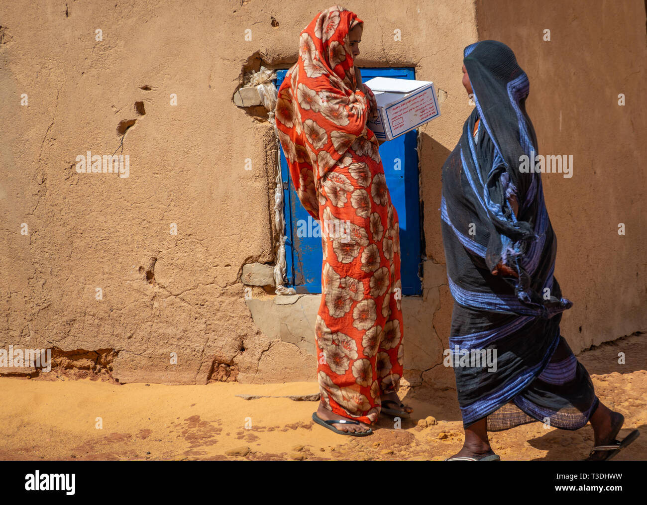 Kerma, Sudan, Februar 10., 2019: Zwei Sudanesische Frauen, eines in einem blauen Kleid, eines in einem roten Kleid, sprechen vor einer Lehmhütte in einer Wüste Dorf. Stockfoto