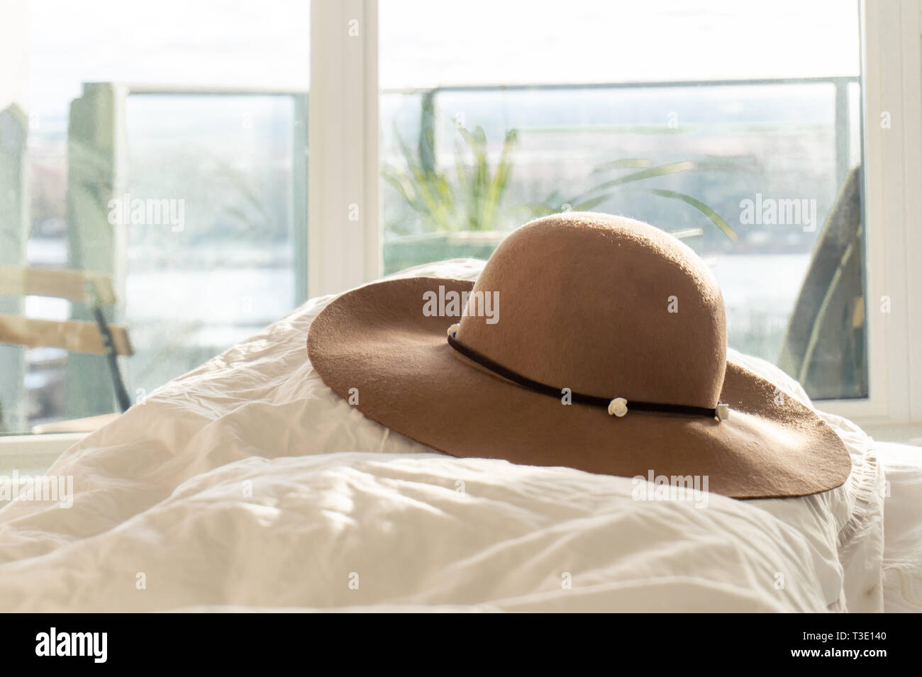 Der Beige Frau runde Sonne hut mit Blumenranken und breiter Krempe, auf einen flauschigen Daunendecke in einem Schlafzimmer, mit Balkon im Hintergrund. Urlaub bildlich darstellen. Stockfoto