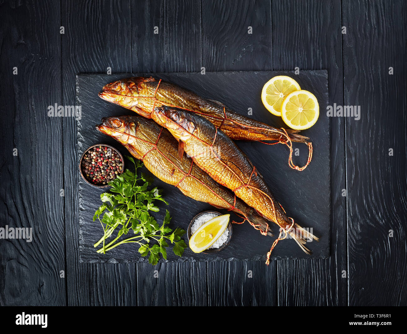 Drei heiß geräucherten Heringe auf schwarzem Schiefer Fach mit Zitronenscheiben, Petersilie und Pfefferkörner, Ansicht von oben, flatlay Stockfoto