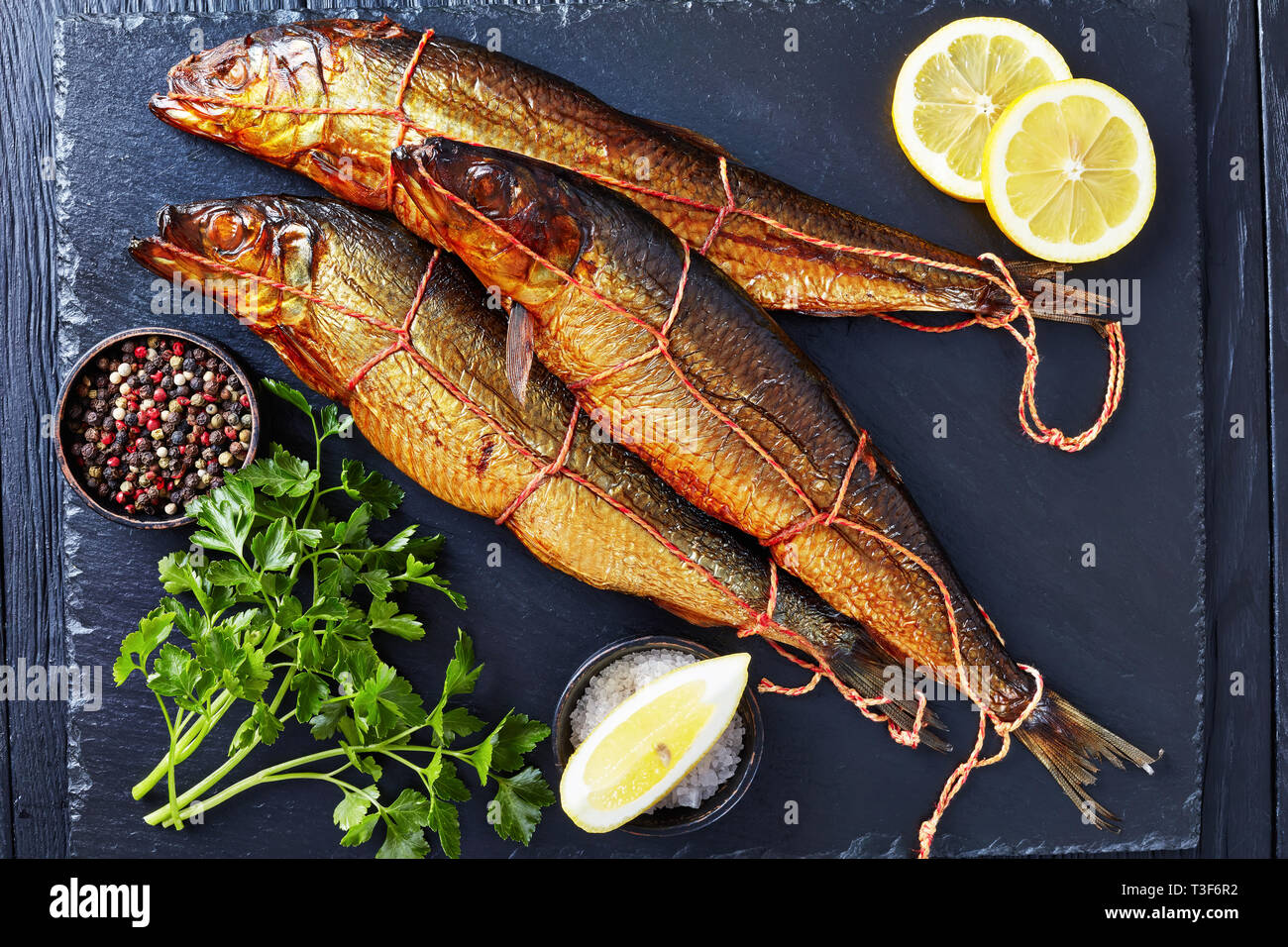 Drei heiß geräucherten Heringe auf schwarzem Schiefer Fach mit Zitronenscheiben, Petersilie und Pfefferkörner, Ansicht von oben, flatlay, close-up Stockfoto