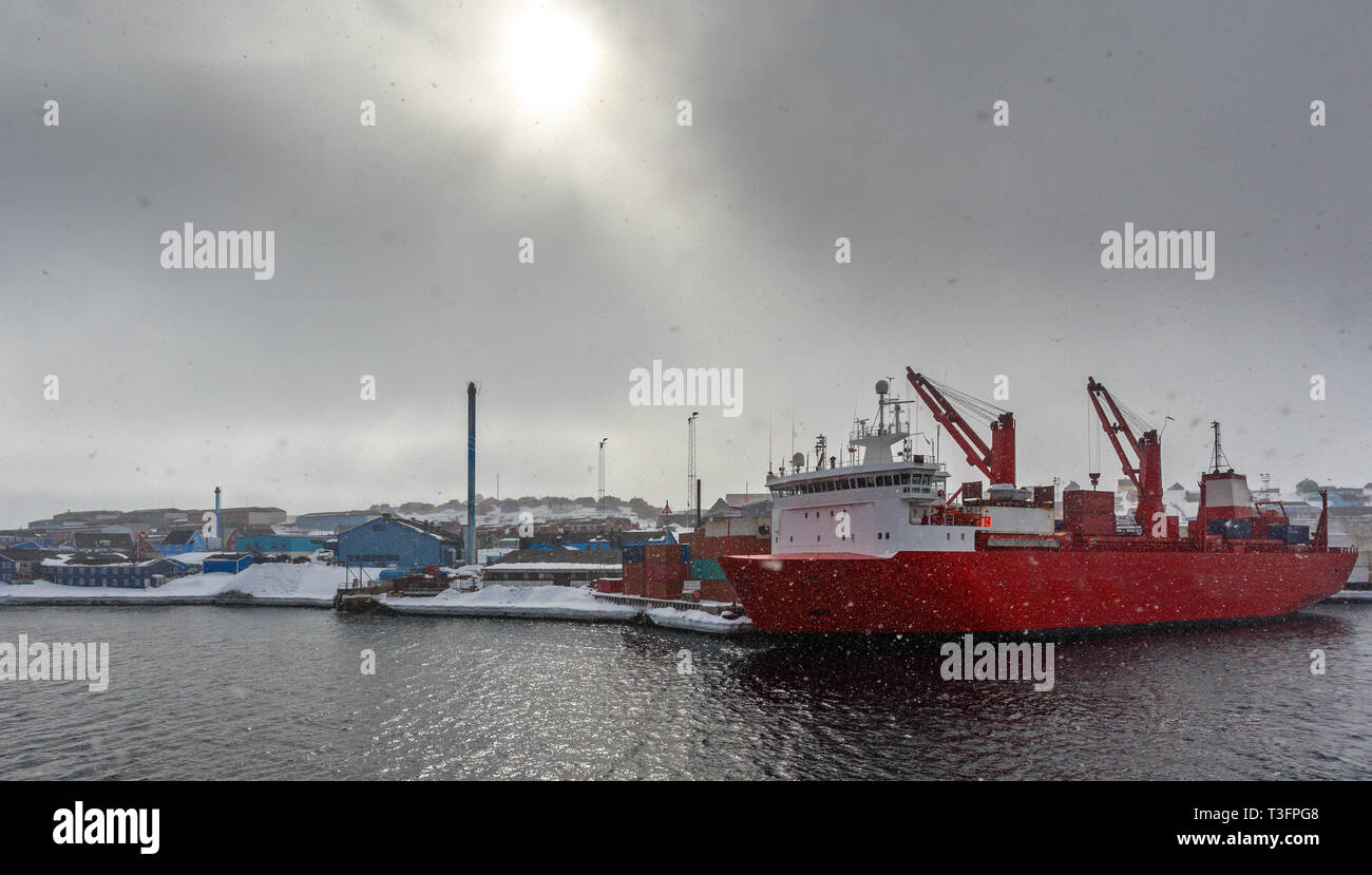 Große rote Frachtschiff unter der Matte Arctic Sun und starker Schneefall im Hafen von Aasiaat, mit Dorf im Hintergrund, Grönland Stockfoto