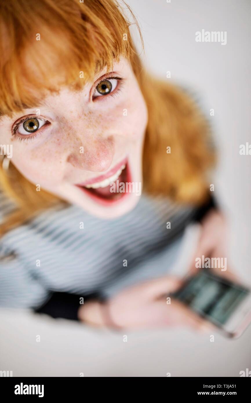 Mädchen, Teenager, Rothaarige mit Smartphone in der Hand, schaut fröhlich, in der Kamera begeistert, studio Shot, Deutschland Stockfoto