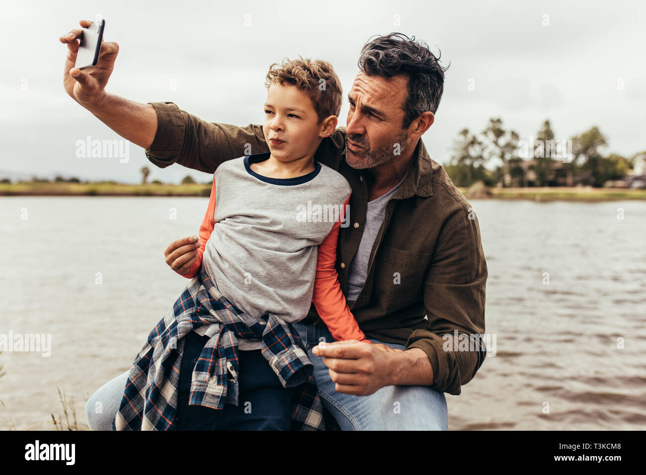 Nahaufnahme von Vater und Sohn eine selfie sitzen in der Nähe von einem See. Glückliches Kind sitzt auf dem Schoß seines Vaters Blick auf das Mobiltelefon, während seine fathe Stockfoto