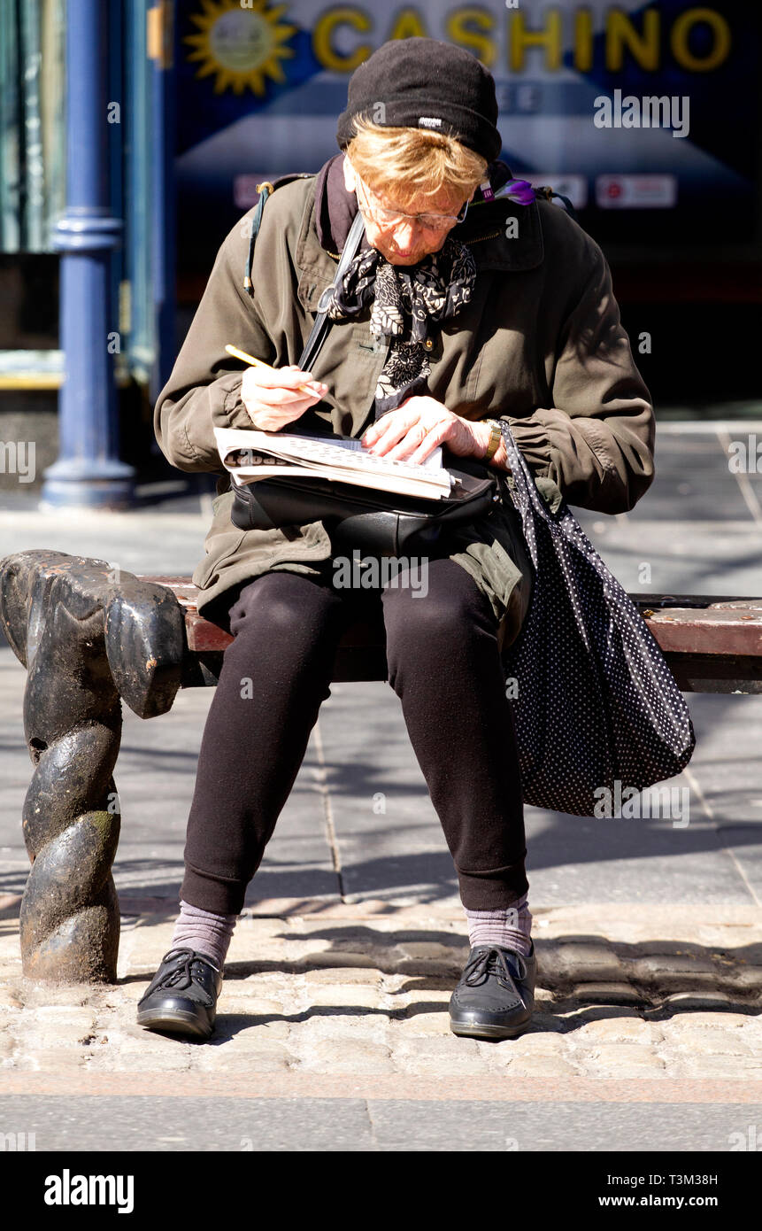 Eine ältere Frau sitzt auf einem Sitz in der Sonne tun die Zeitung Kreuzworträtsel in Dundee, Großbritannien Stockfoto