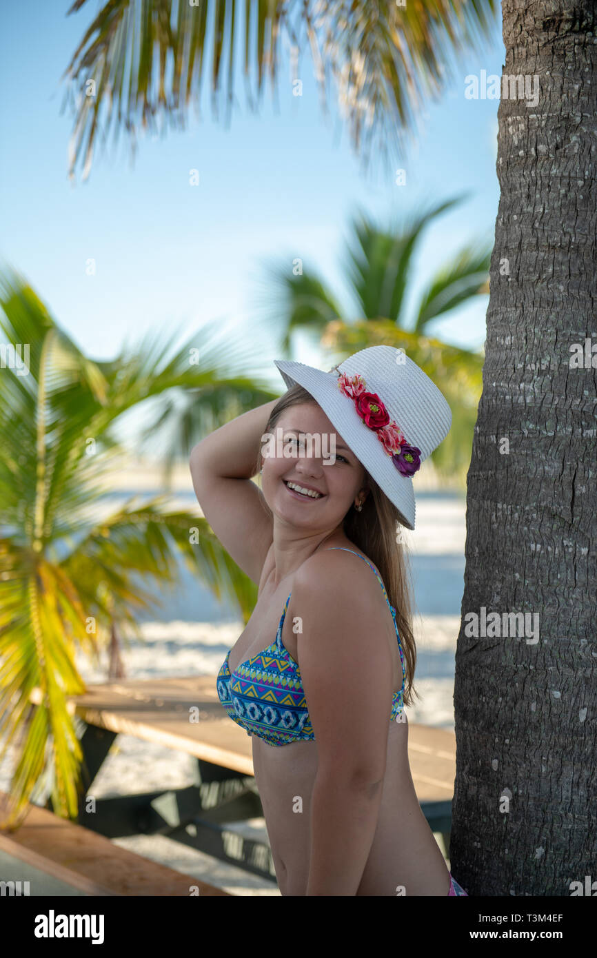 Junge Frau im Bikini mit Hut stehend in der Nähe einer Palme am Strand und posieren. Sonnenbaden und Solarium in den USA Stockfoto