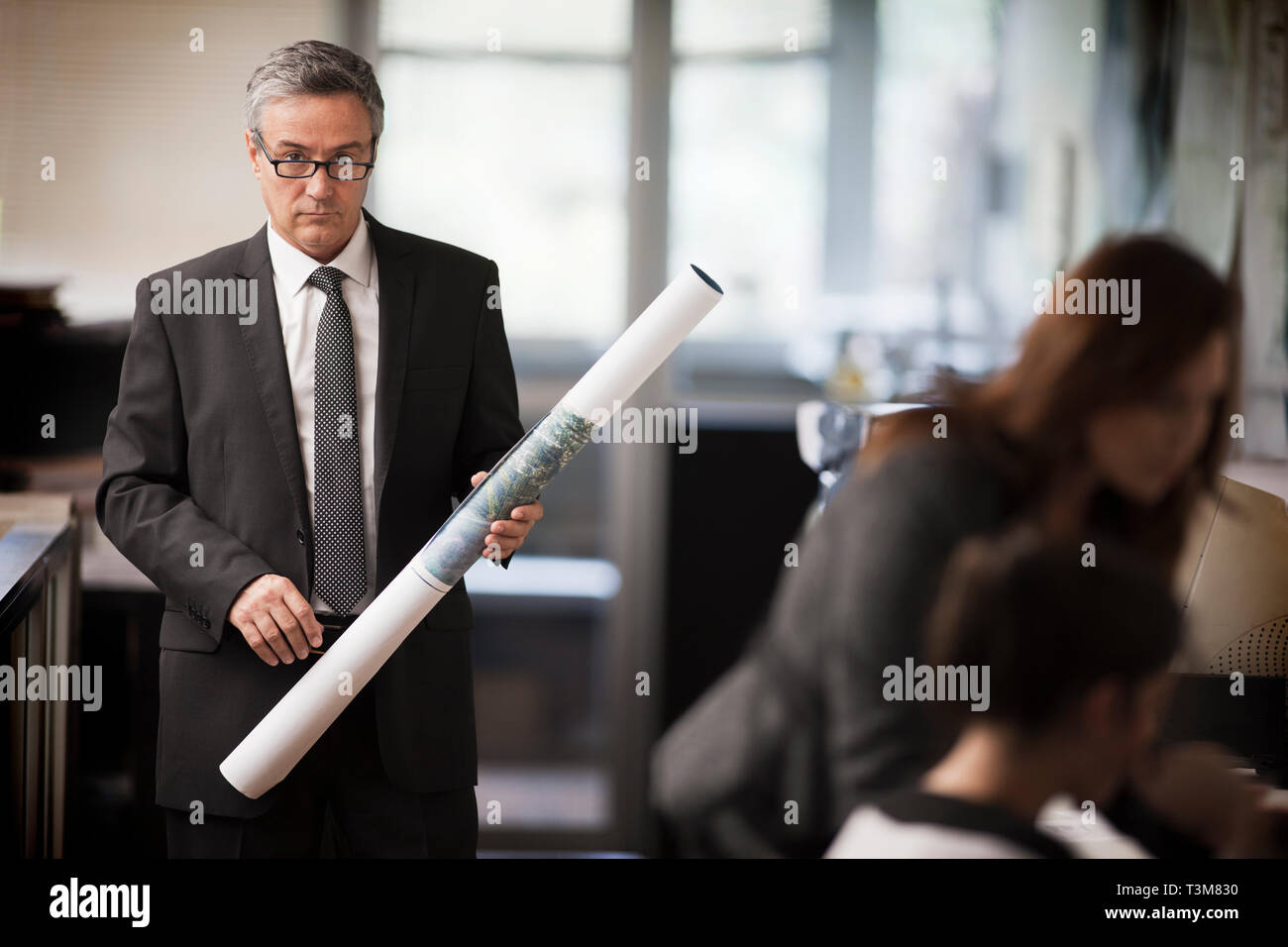 Reifen Geschäftsmann Holding eine Rolle Papier schaut über seine Brille, während für ein Portrait posiert, wie er für zwei weiblichen Kollegen wartet. Stockfoto