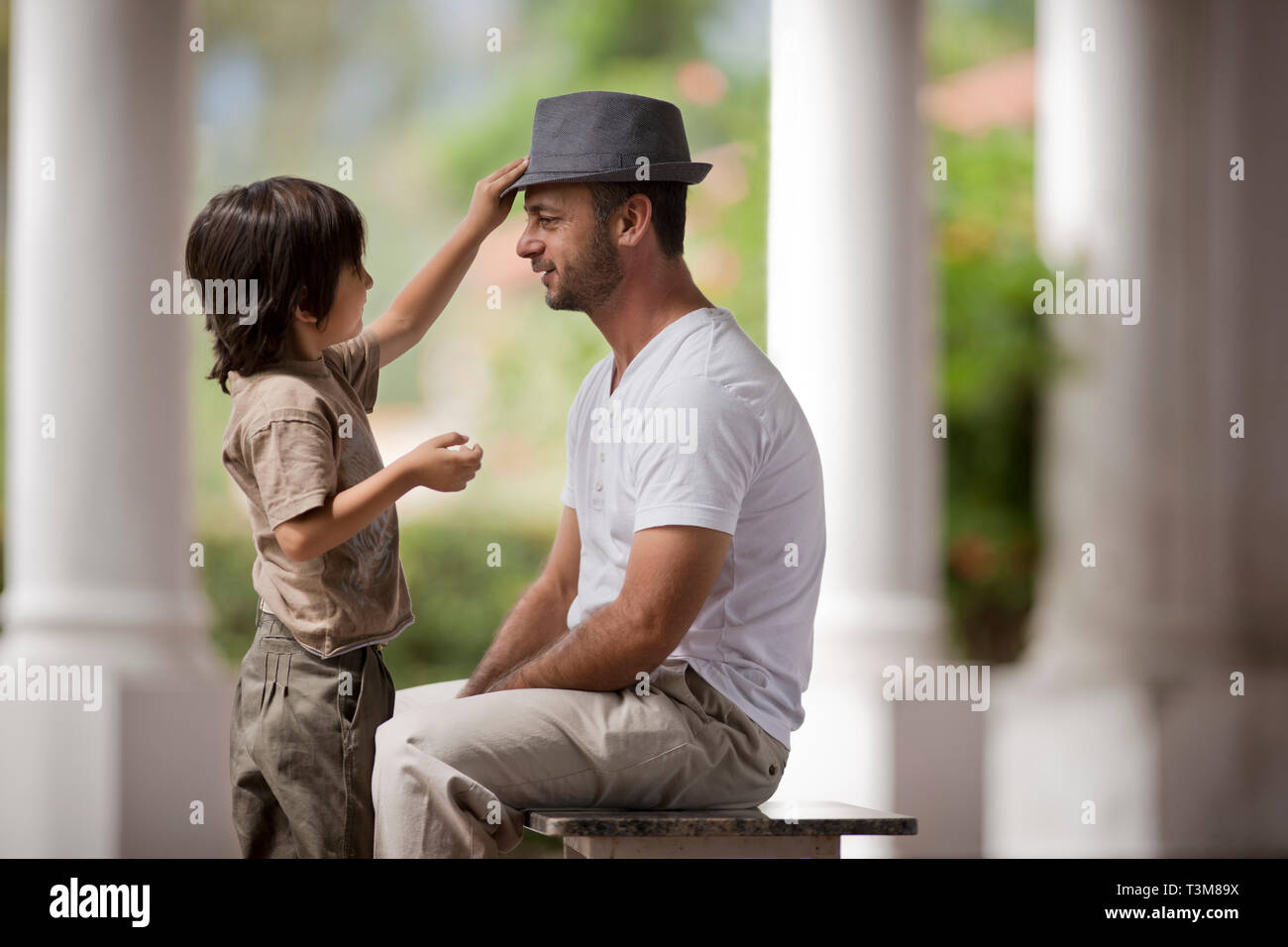Thirtysomething Mann sitzt als sein junger Sohn spielt mit seinem Trilby Hut. Stockfoto