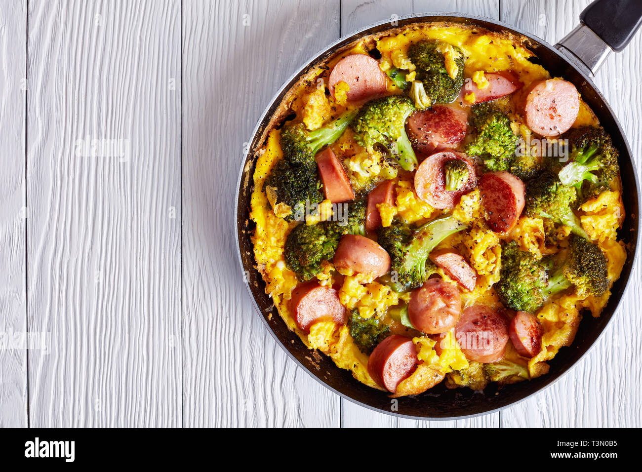 Leckere heiße Omelette mit Brokkoli und Würste in einer Pfanne auf einem weißen Holztisch, horizontale Ansicht von oben, flach, Nahaufnahme, Kopie Raum Stockfoto