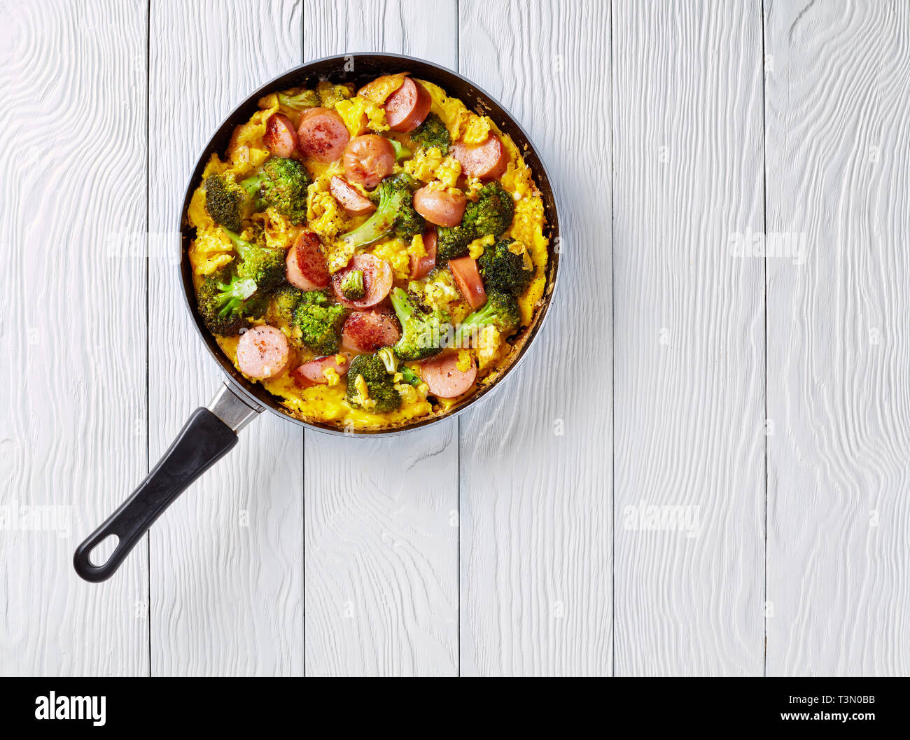 Leckere heiße Omelette mit Brokkoli und Würste in einer Pfanne auf einem weißen Holztisch, horizontale Ansicht von oben, flach, leeren Raum rechts Stockfoto
