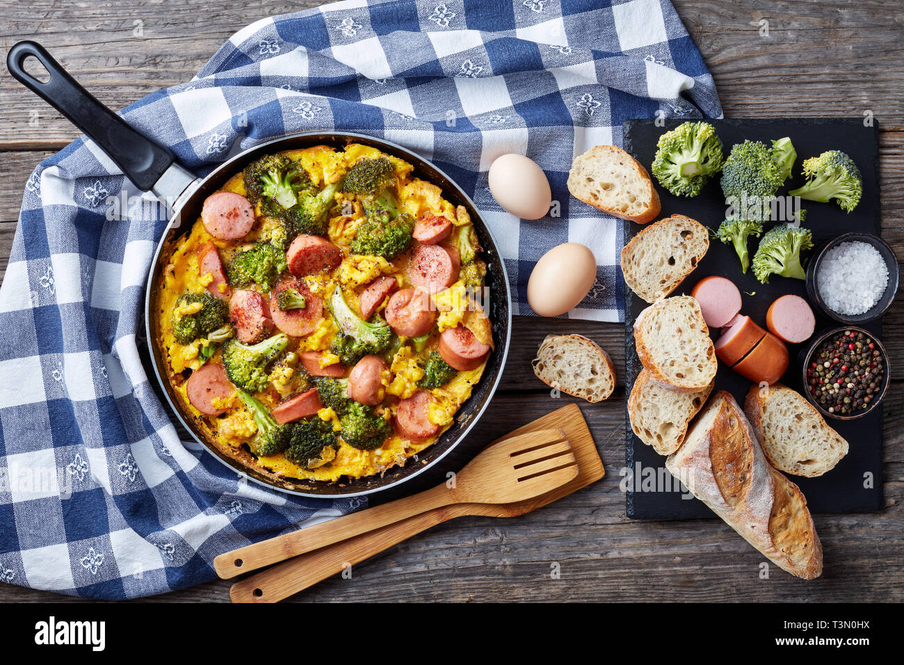 Leckere heiße Omelette mit Brokkoli und Würste in einer Pfanne auf einem alten Holztisch mit Zutaten in Scheiben geschnitten und knusprigem Baguette Vollkorn, Stockfoto