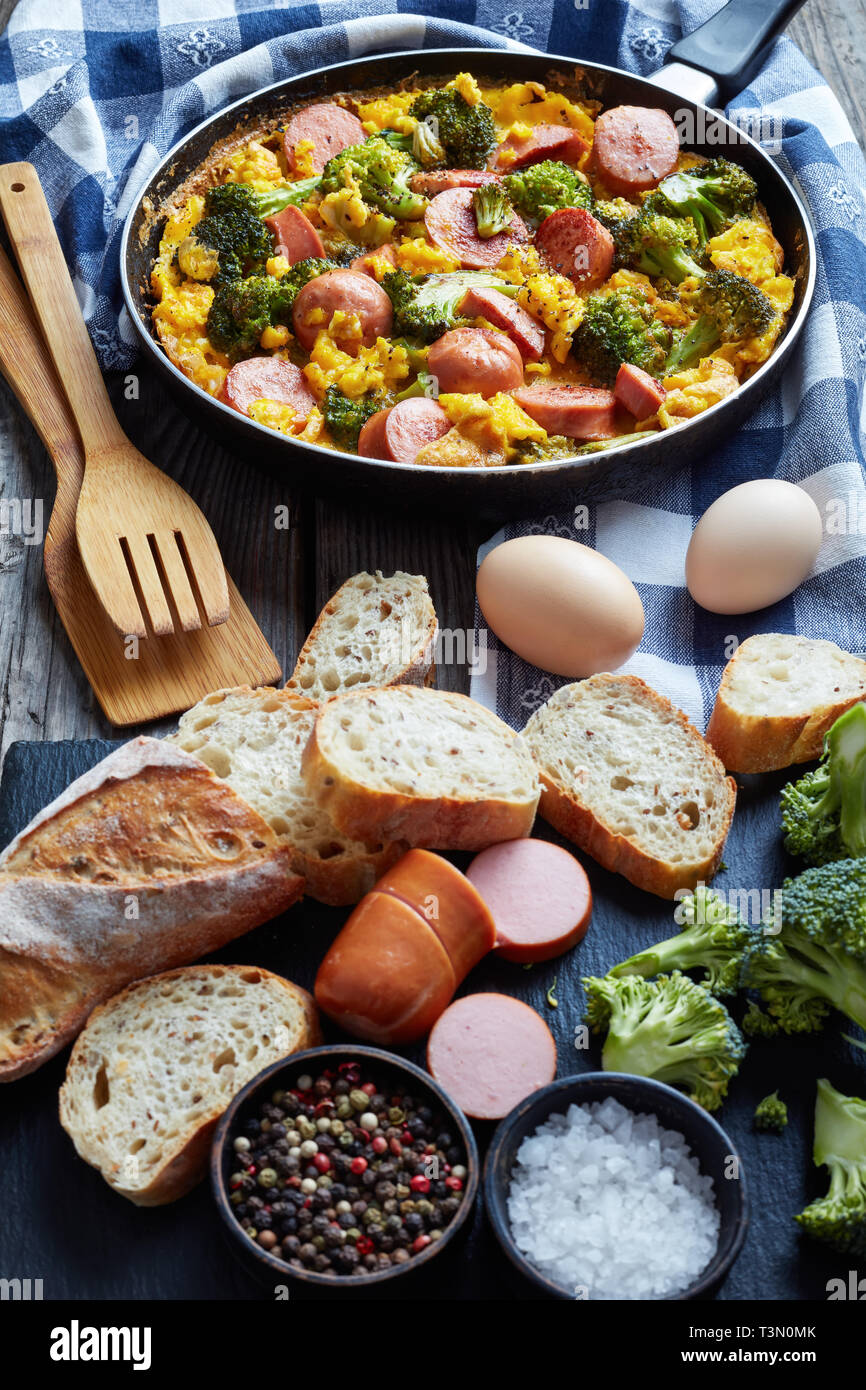Köstliche warme Rühreier mit Brokkoli und Würste in einer Pfanne auf einem alten Holztisch mit Zutaten in Scheiben geschnitten und crusty Vollkorn Französische bagu Stockfoto