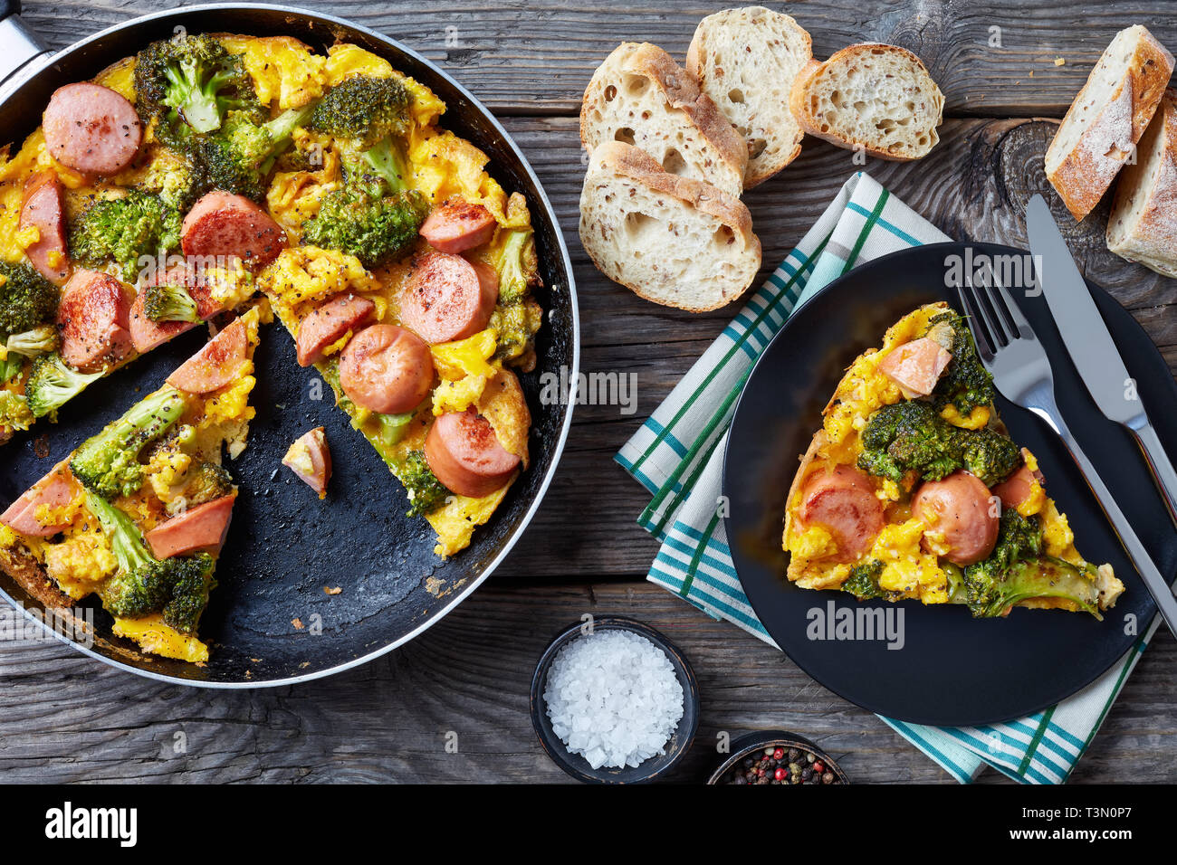 Omelette mit Brokkoli und Würste in einer Pfanne und auf Teller serviert auf einem alten grauen rustikalen Holztisch mit in Scheiben geschnittenen crusty Vollkorn Französisches baguett Stockfoto