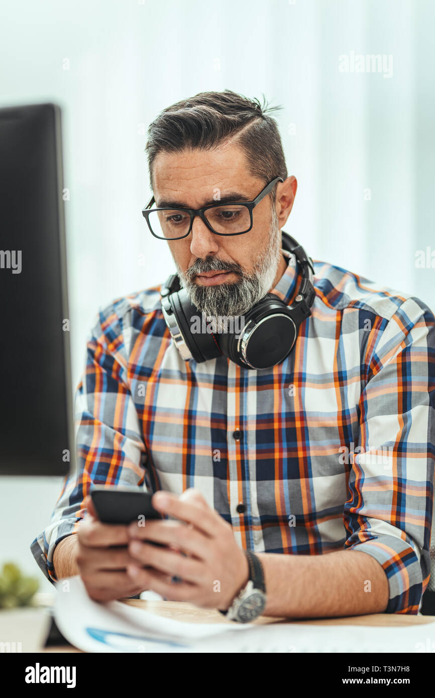 Ein erfolgreicher Unternehmer, mit Kopfhörer um den Hals, ist das Schreiben von Nachrichten, die auf einem Smartphone im Büro. Stockfoto