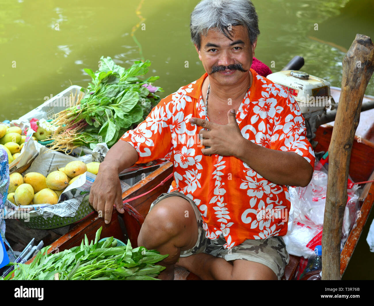 Fröhlichen Thai boot Anbieter verkauft frisches grünes Gemüse und süßen Mangos von seinem Boot und posiert für die Kamera. Stockfoto