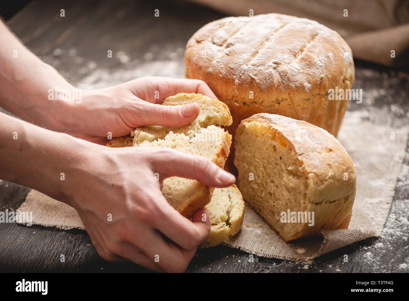 Frauen Hände brechen hausgemachten Naturprodukten frisches Brot mit einer goldenen Kruste auf eine Serviette auf einem alten Holz- Hintergrund. Das Konzept der Backen Backwaren Stockfoto