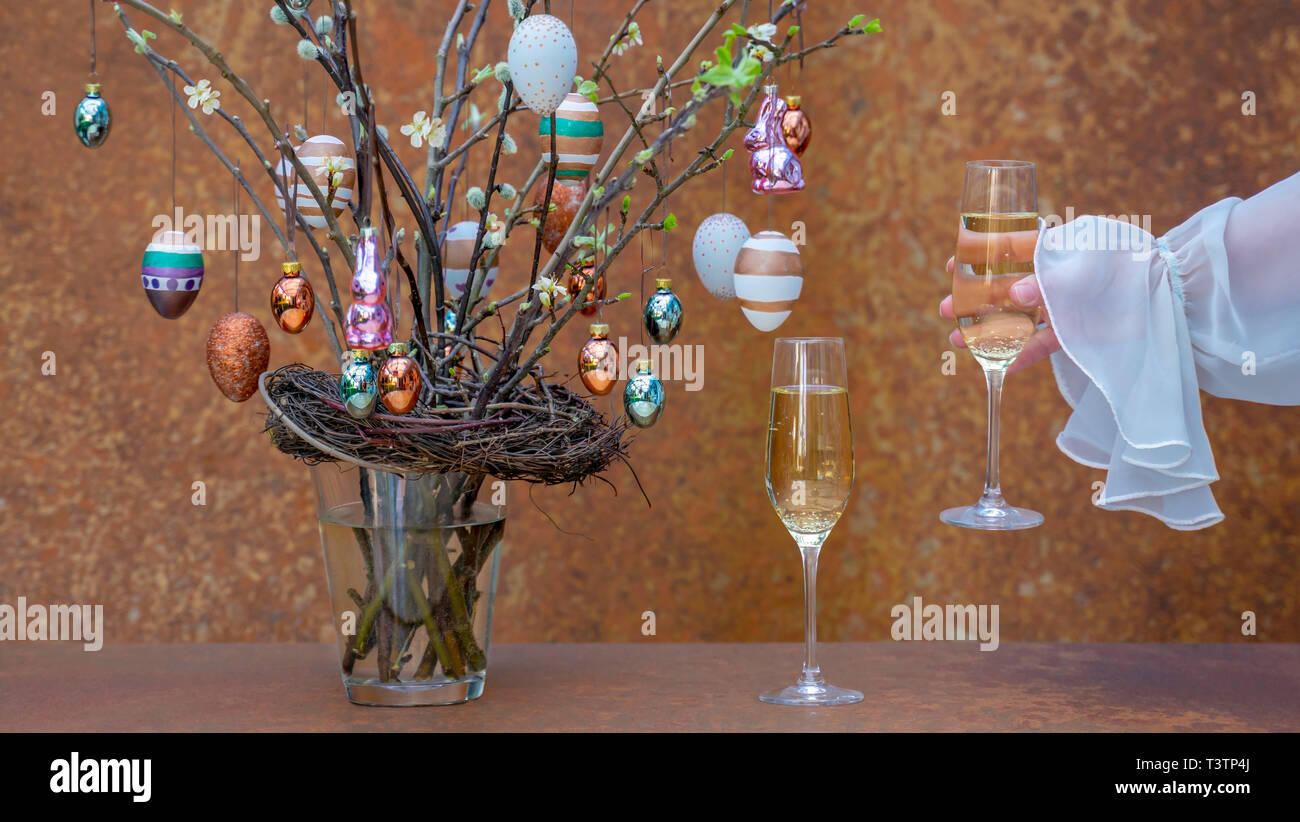 Branchen mit Blüten und Knospen. Ostereier und Osterhasen hängen an den Zweigen. Womans hand Champagner Glas. Festliche Kleidung. Stockfoto