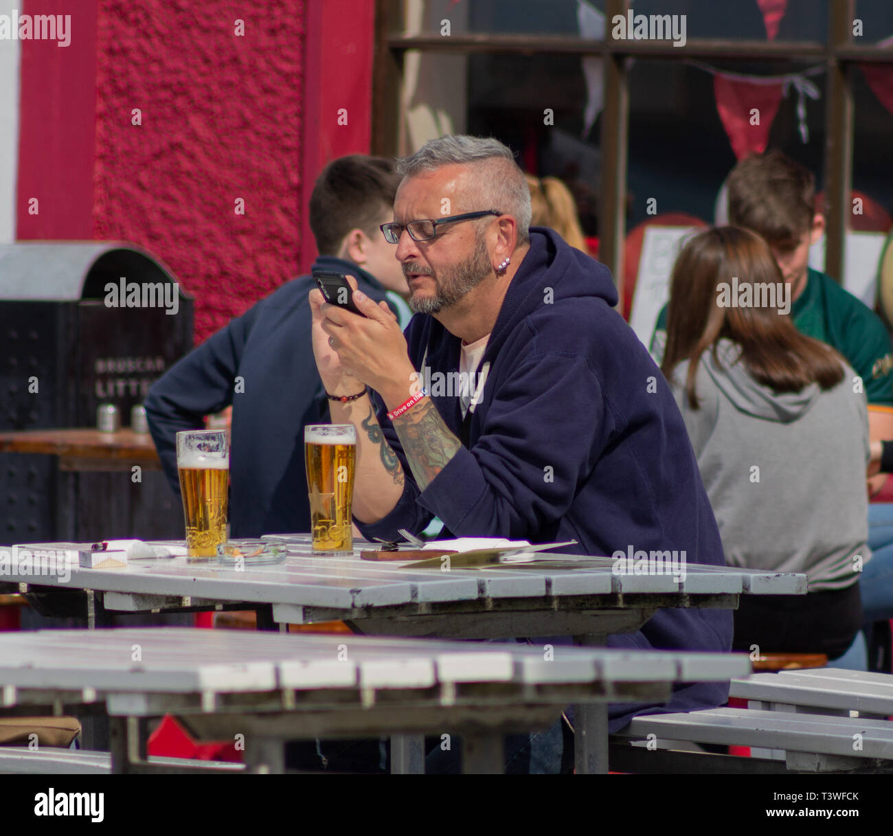 Mittleres alter Mann mit Handy außerhalb ein Pub mit Gläser Bier auf seinem Tisch. Stockfoto