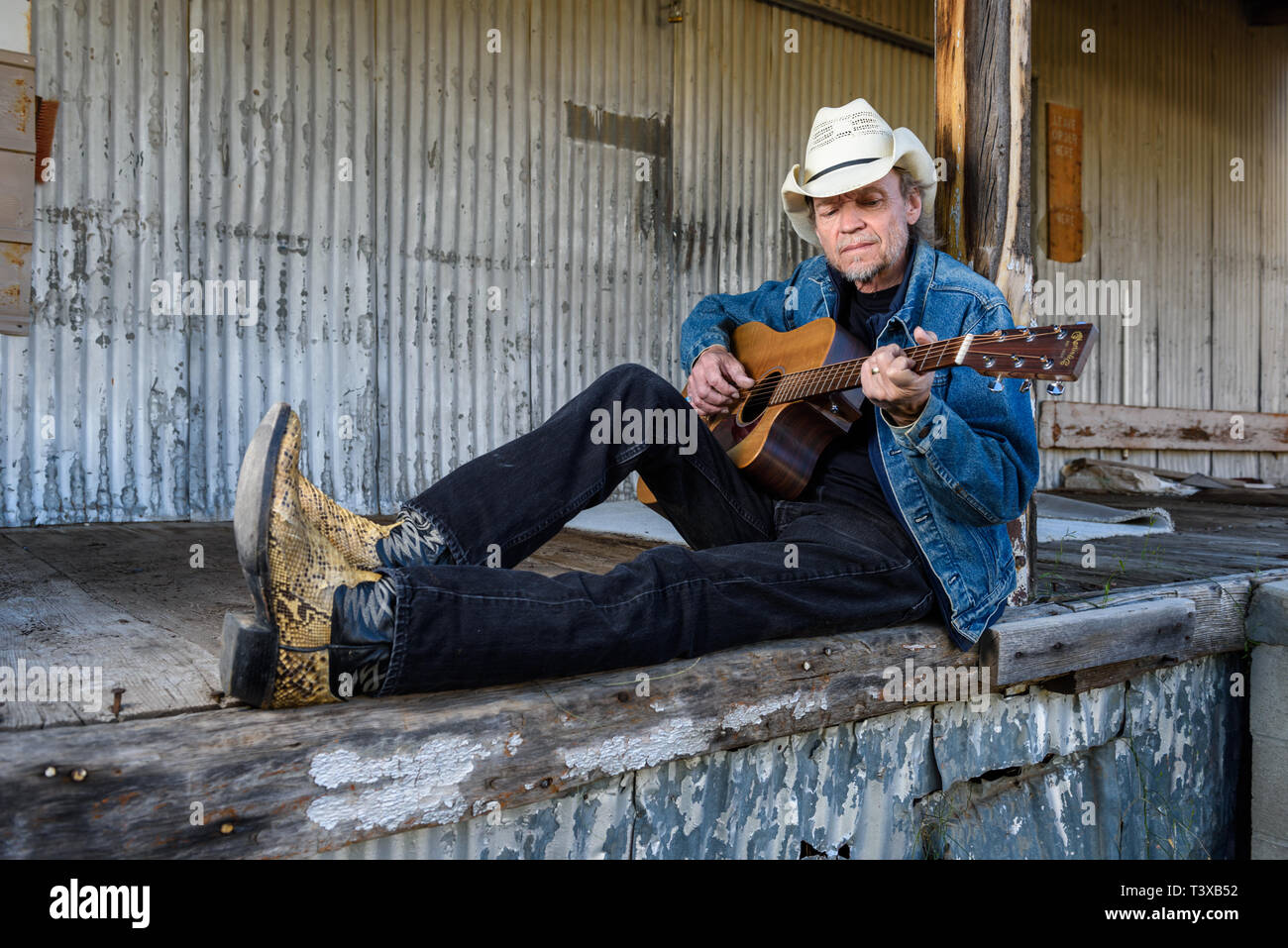 Alte Cowboy trägt Cowboyhut und Stiefel spielt eine Gitarre beim Sitzen auf einem alten Dock. Stockfoto