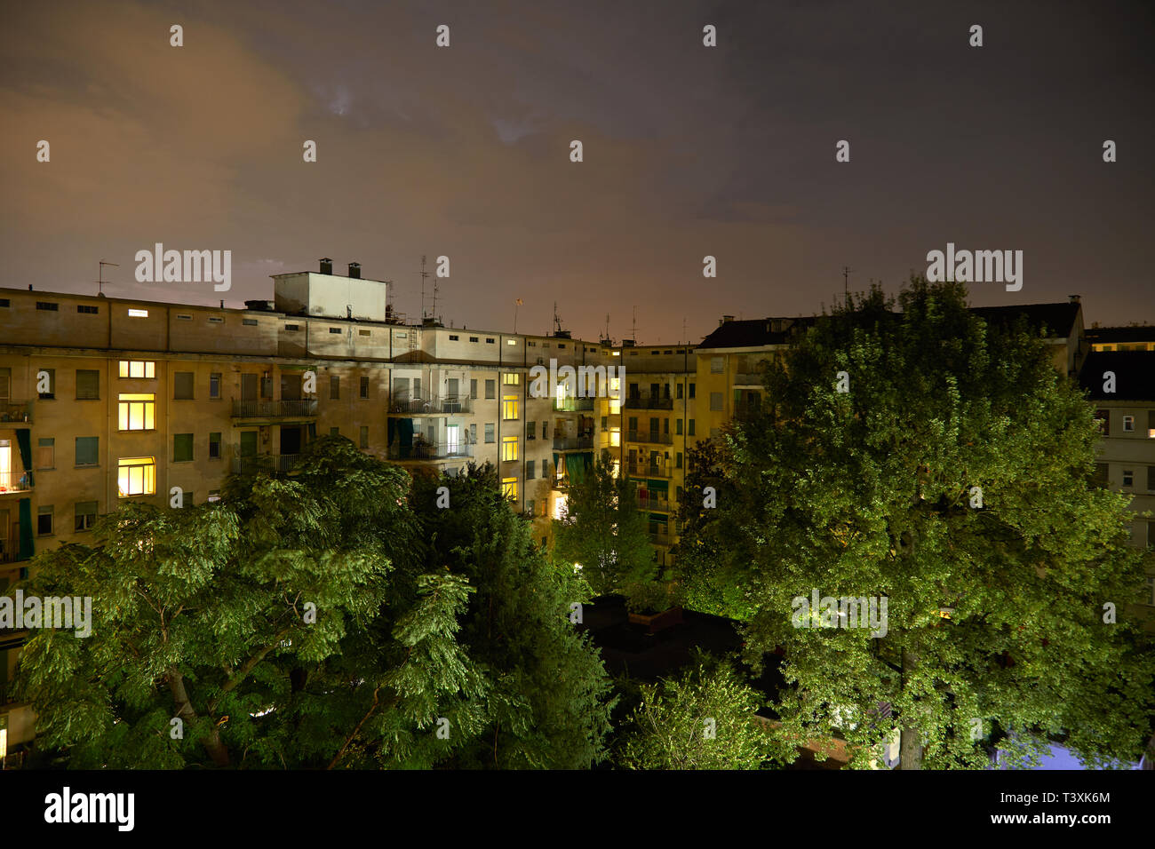 Gebäude und grüne Bäume bei Nacht, beleuchtete bewölkter Himmel Stockfoto