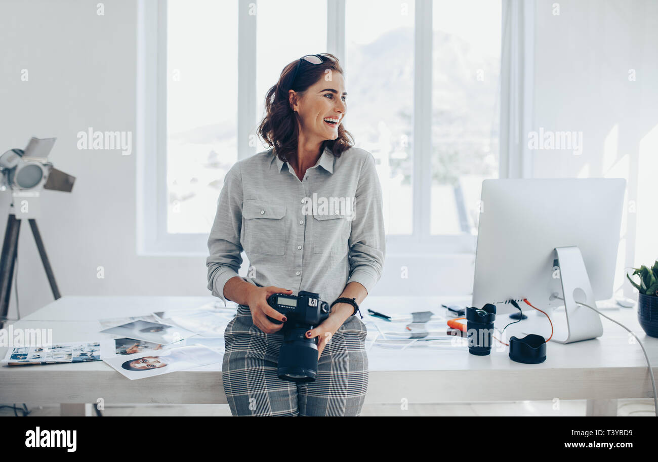 Fröhliche Frau Fotograf mit einer professionellen Kamera. Weibliche in Casuals an den Schreibtisch mit digitaler Kamera in der Hand lehnte und Wegsehen lachen. Stockfoto