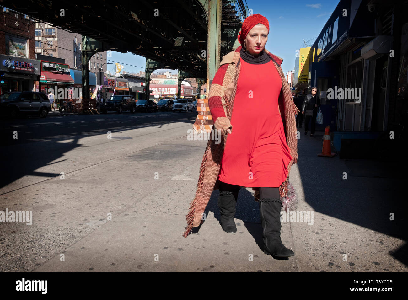 Eine attraktive Frau in Rot trägt einen langen Mantel und Stiefel Spaziergänge unter der El auf Roosevelt Avenue in Jackson Heights, Queens, New York City. Stockfoto