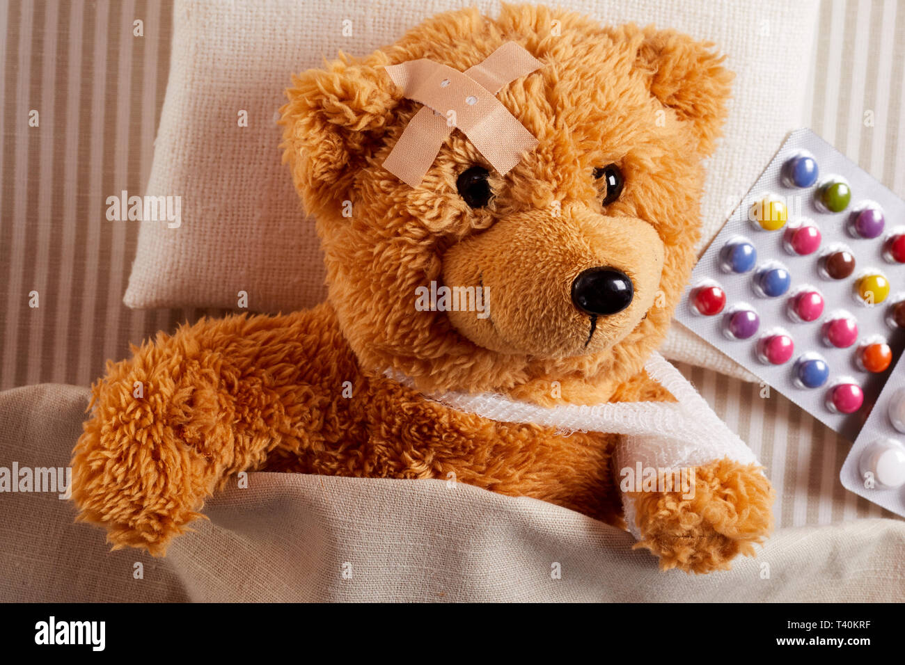 Wenig verletzte Teddybär lag krank im Bett mit einem Pflaster auf der Stirn  und bandagierten Arm in einer Schlinge neben einer Blisterpackung von  bunten Pillen in einem Stockfotografie - Alamy