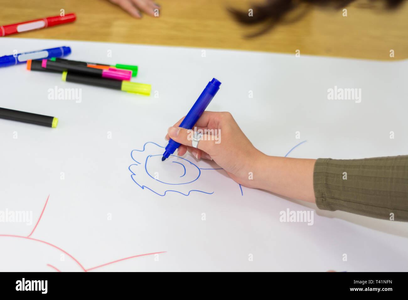 Student hand mit blauem Filzstift zeichnen Einfache stickman auf großen weißen Papier auf dem Schreibtisch im Klassenzimmer. Bunte Kugelschreiber auf Papier verschüttet. Bildung Konzept Stockfoto