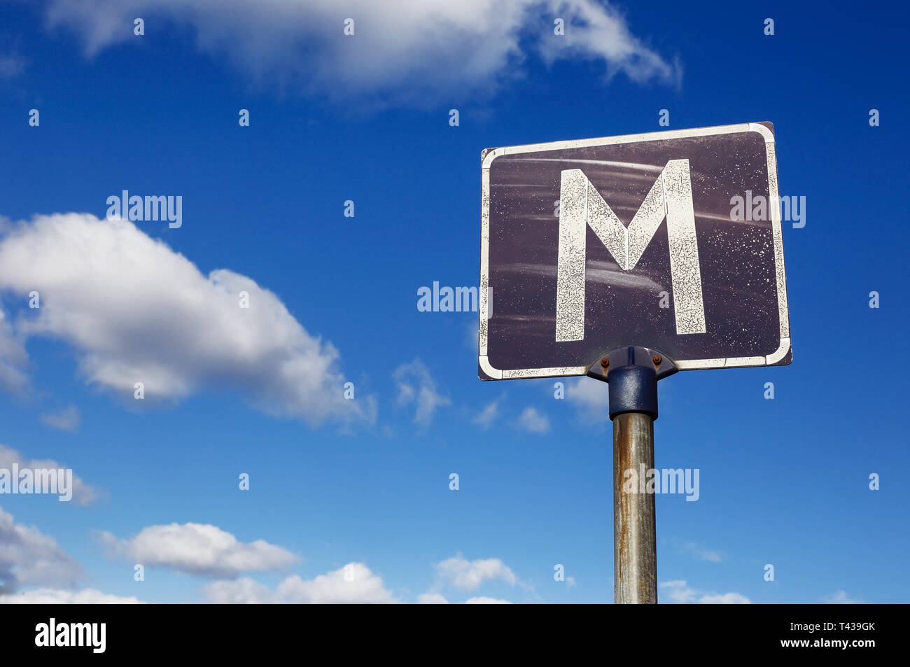 Eine verwitterte Schild mit dem Buchstaben M zeigen einen Treffpunkt auf einer schmalen Straße vor einem blauen bewölkten Himmel. Stockfoto