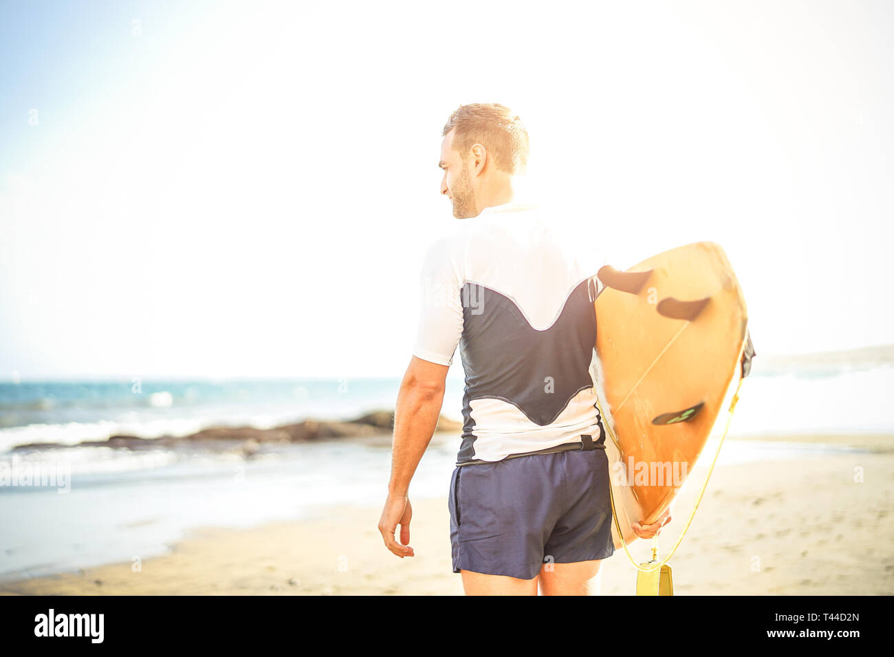 Junge surfer Holding sein Surfbrett auf die Wellen zum Surfen - schöner Mann stehen auf dem Strand bei Sonnenuntergang Training zu surfen. Stockfoto