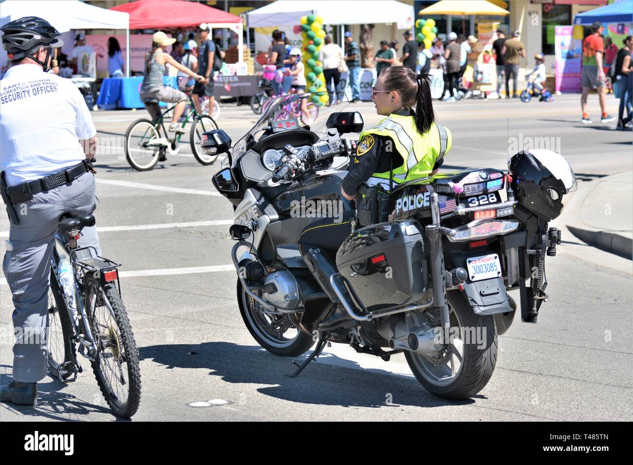 Eine junge Polizistin mit einem langen Pony-Schwanz auf dem Fahrrad bei einer öffentlichen Veranstaltung, eine faltbare M-16 auf dem Rücken mit ihrem vollen Helm Stockfoto