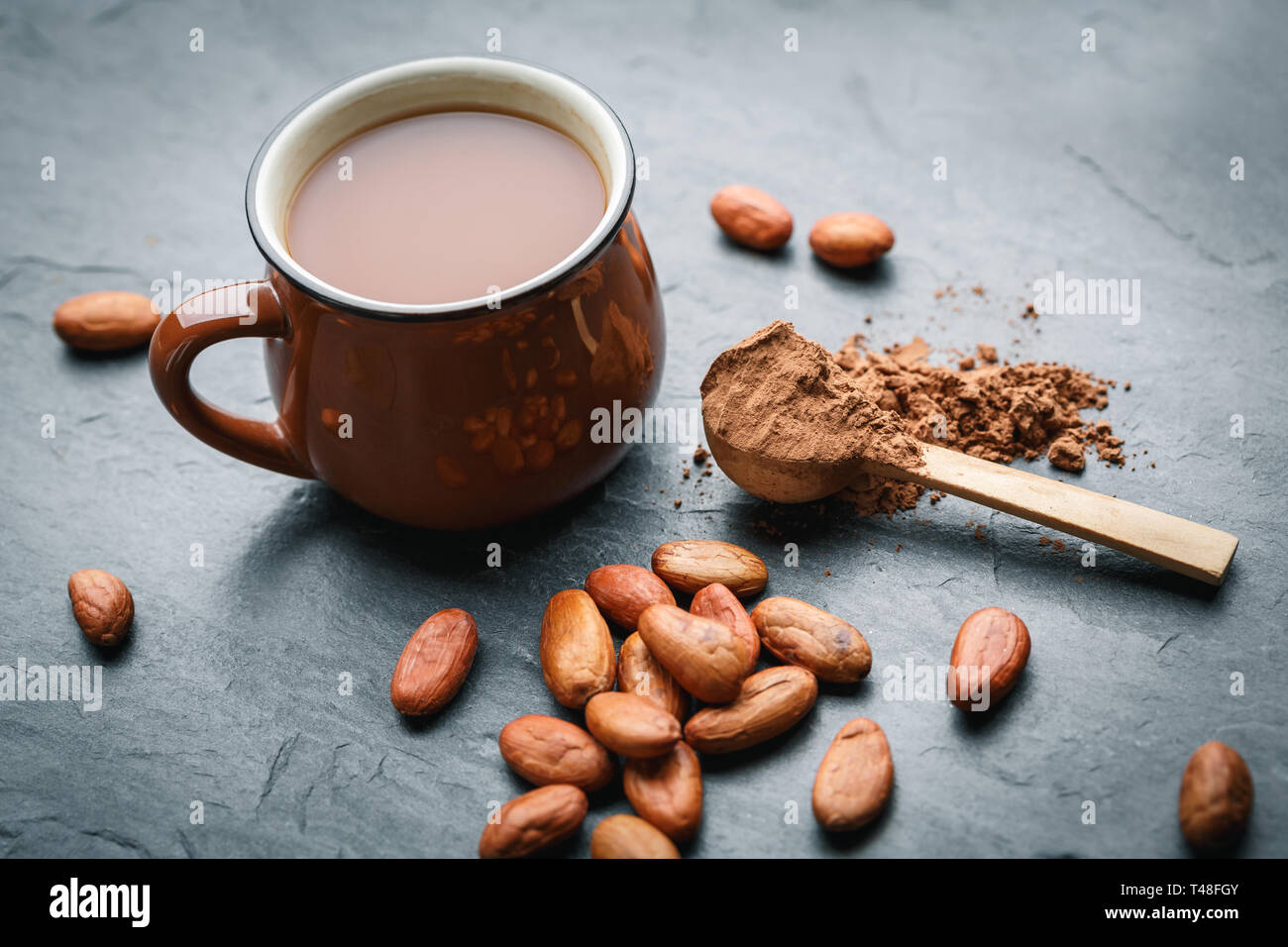 Braune Tasse Kakao trinken mit Kakaobohnen und Pulver in kleinen Holzlöffel auf schwarzen Stein Hintergrund Stockfoto