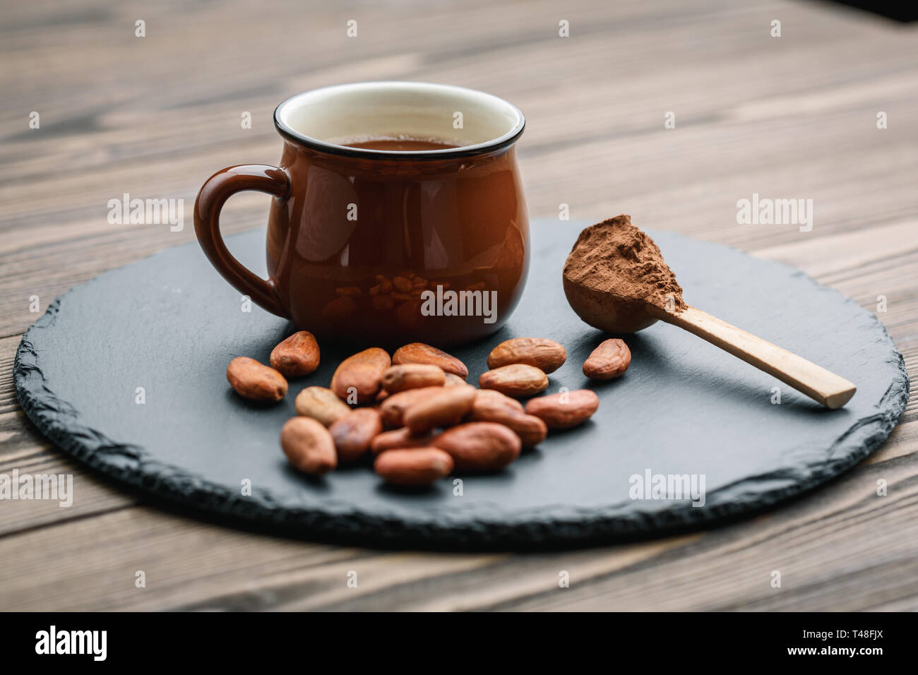 Braune Tasse Kakao trinken mit Kakaobohnen und Pulver in kleinen Holzlöffel auf schwarzen Stein Hintergrund Stockfoto