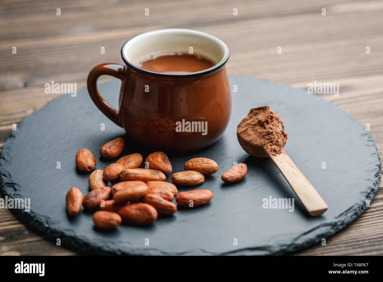 Braune Tasse Kakao trinken mit Kakaobohnen und Pulver in kleinen Holzlöffel auf schwarzen Stein Hintergrund Stockfoto