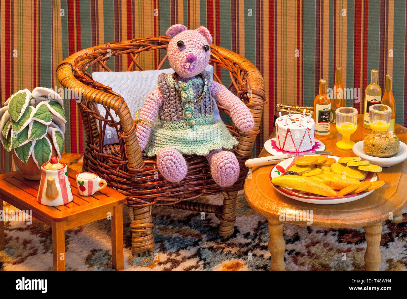 Handgefertigter Häkelbär sitzt in einem gemütlichen Puppenhaus-Wohnzimmer und genießt Tee, Kuchen und Gebäck neben einem Korbstuhl und farbenfrohen Vintage-Dekor. Stockfoto