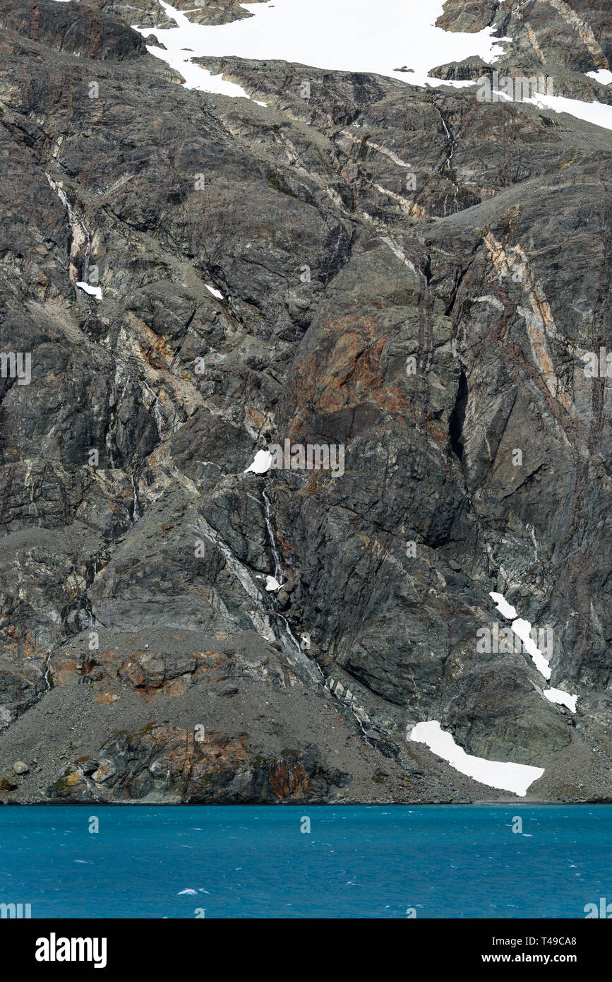 Verwitterten grauen Felswand mit Orange und Weiß mineral Venen, Schnee, Patches, und glazialen blaues Wasser, Drygalski Fjord, Südgeorgien Stockfoto