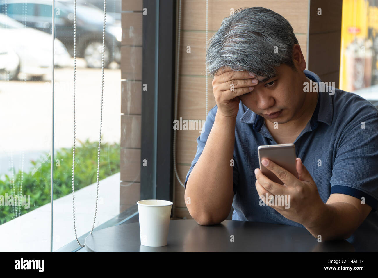 Asiatische Männer mittleren Alters (40 Jahre alt), gestresst und müde und mit Smartphone, Sitzen in schnell Restaurant. Auf dem Tisch ist ein White Paper Cup Stockfoto