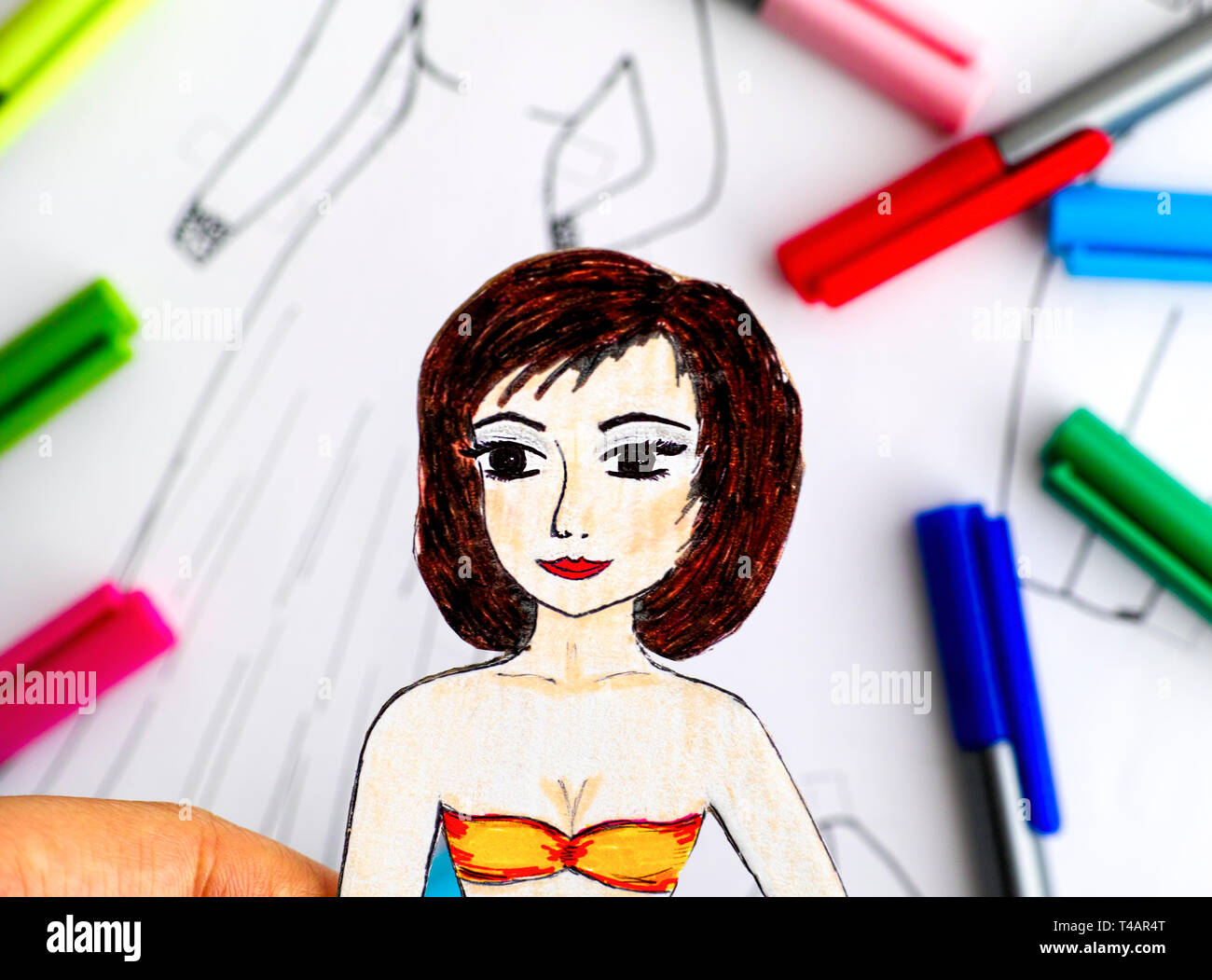 Paper doll in der menschlichen Hand gegen Papier mit Zeichnung Kleidung und Farbstifte. Close-up. Stockfoto Paper doll in der menschlichen Hand gegen Papier mit Zeichnung Kleidung und Farbstifte. Close-up. Stockfoto