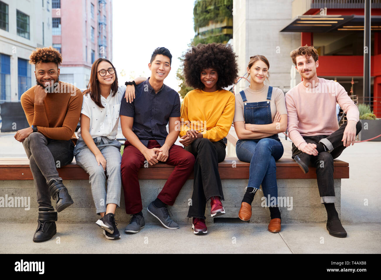 Sechs junge erwachsene Freunde in einer Reihe sitzen auf einer Bank in einer Stadtstraße lächelnd in die Kamera, volle Länge Stockfoto
