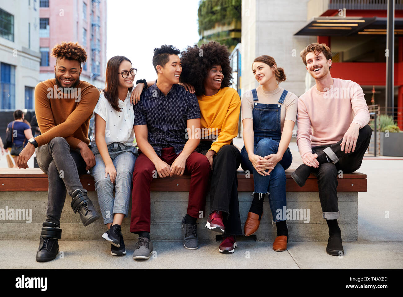 Sechs junge erwachsene Freunde in einer Reihe sitzen auf einer Bank in der Straße sich einander, volle Länge Stockfoto