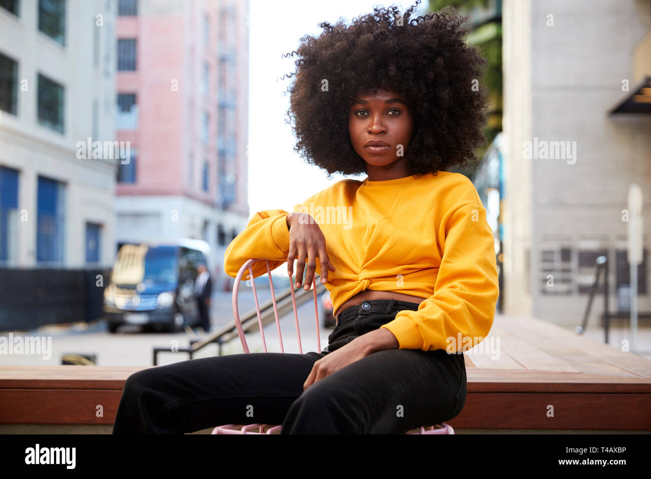 Junge schwarze Frau mit afro Haar und gelb Top sitzt auf einem Stuhl in der Straße an der Kamera auf der Suche Stockfoto