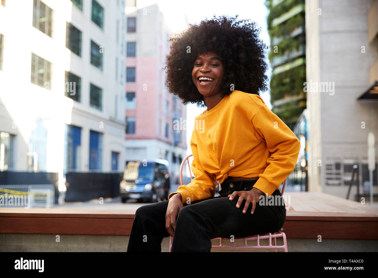 Junge schwarze Frau mit afro Haar sitzt auf einem Stuhl in der Straße an der Kamera lachend Stockfoto