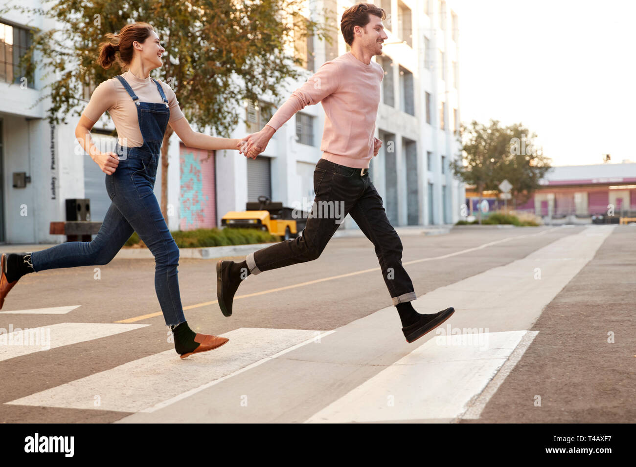 Tausendjährige hipster Paar laufen über die Straße in der Stadt halten sich an den Händen, Seitenansicht Stockfoto
