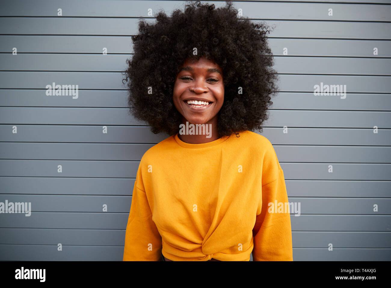 Junge schwarze Frau mit Afro stehend gegen Grau Sicherheit Rollläden, Lächeln für die Kamera, in der Nähe Stockfoto
