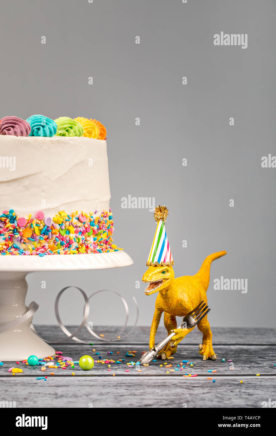 Hungrig Spielzeug Dinosaurier einen Hut tragen und halten eine Gabel neben einer Geburtstagstorte auf grauem Hintergrund Stockfoto