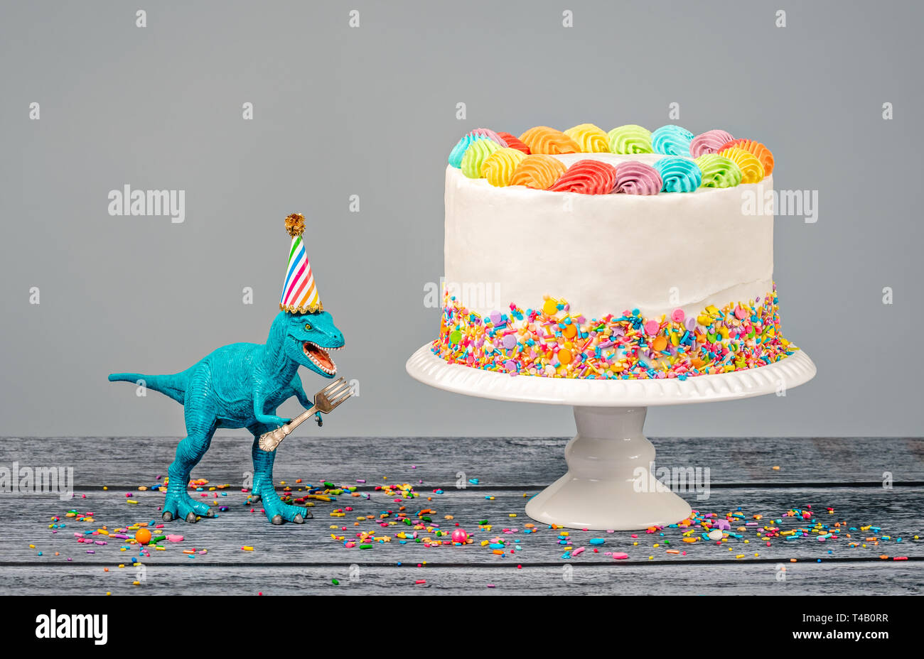 Hungrig Spielzeug Dinosaurier einen Hut tragen und halten eine Gabel neben einer Geburtstagstorte auf grauem Hintergrund Stockfoto