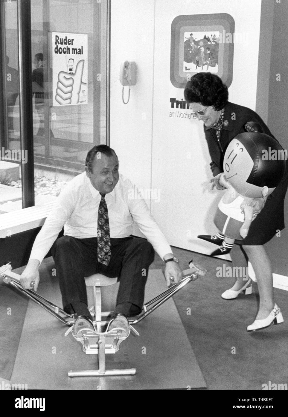 Bundesinnenminister Hans Dietrich Genscher (FDP) probiert am 24.05.1972 im Neueröffneten "Haus des Deutschen Sports in Frankfurt am Main ein Rudergerät aus. Neben ihm steht Annemarie Griesinger, die baden-württembergische Ministerin für gesellschaftspolitische Angelegenheiten. Sie hält ein aufblasbares neues Modell des "Trimmi" in Händen. | Verwendung weltweit Stockfoto