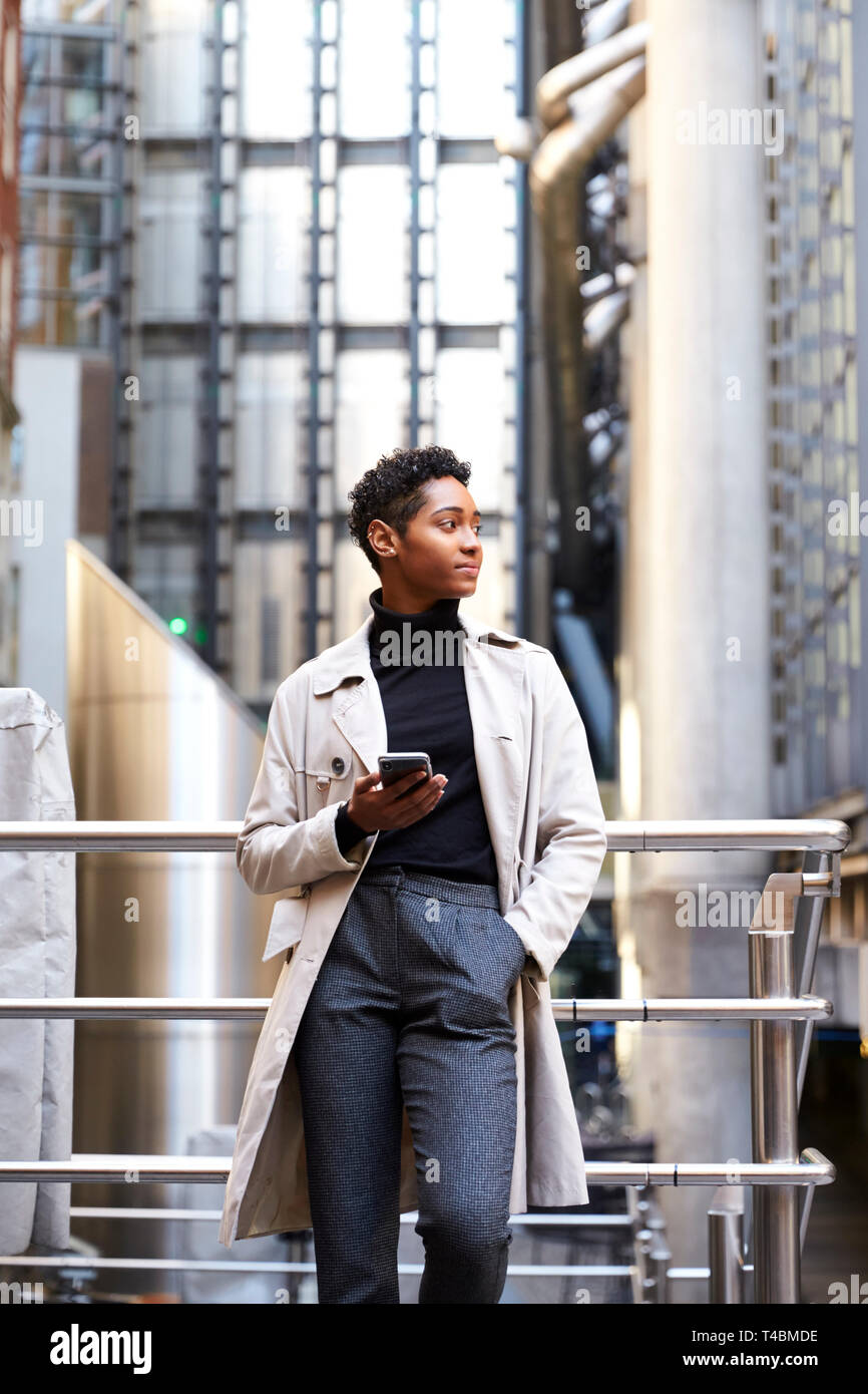 Modische junge schwarze Frau, die in der Stadt auf einem Geländer holding Smartphone, niedrigen Winkel, vertikal Stockfoto